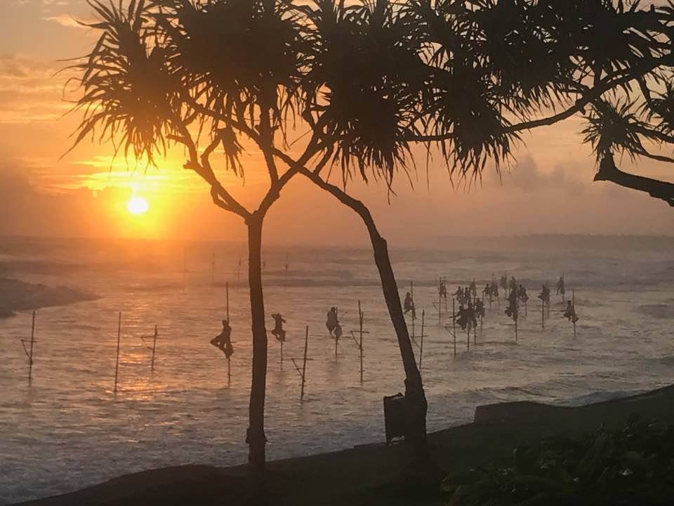 Golden sunset over the ocean with silhouetted stilt fishermen and palm trees, a view near Ko Lake Villa, Sri Lanka.