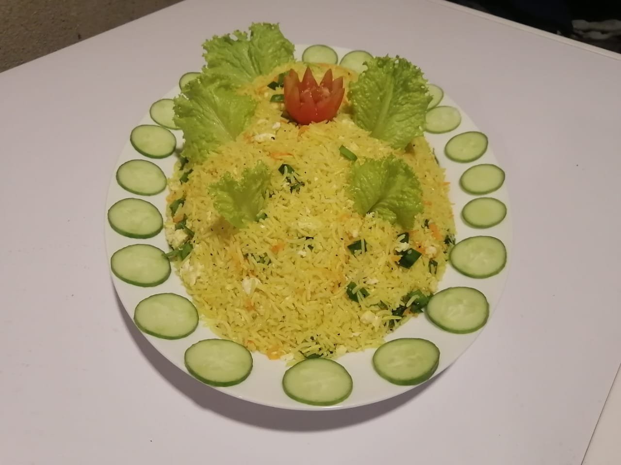 yellow rice dish, garnished with fresh lettuce, cucumber slices, and a tomato flower, on a white plate.