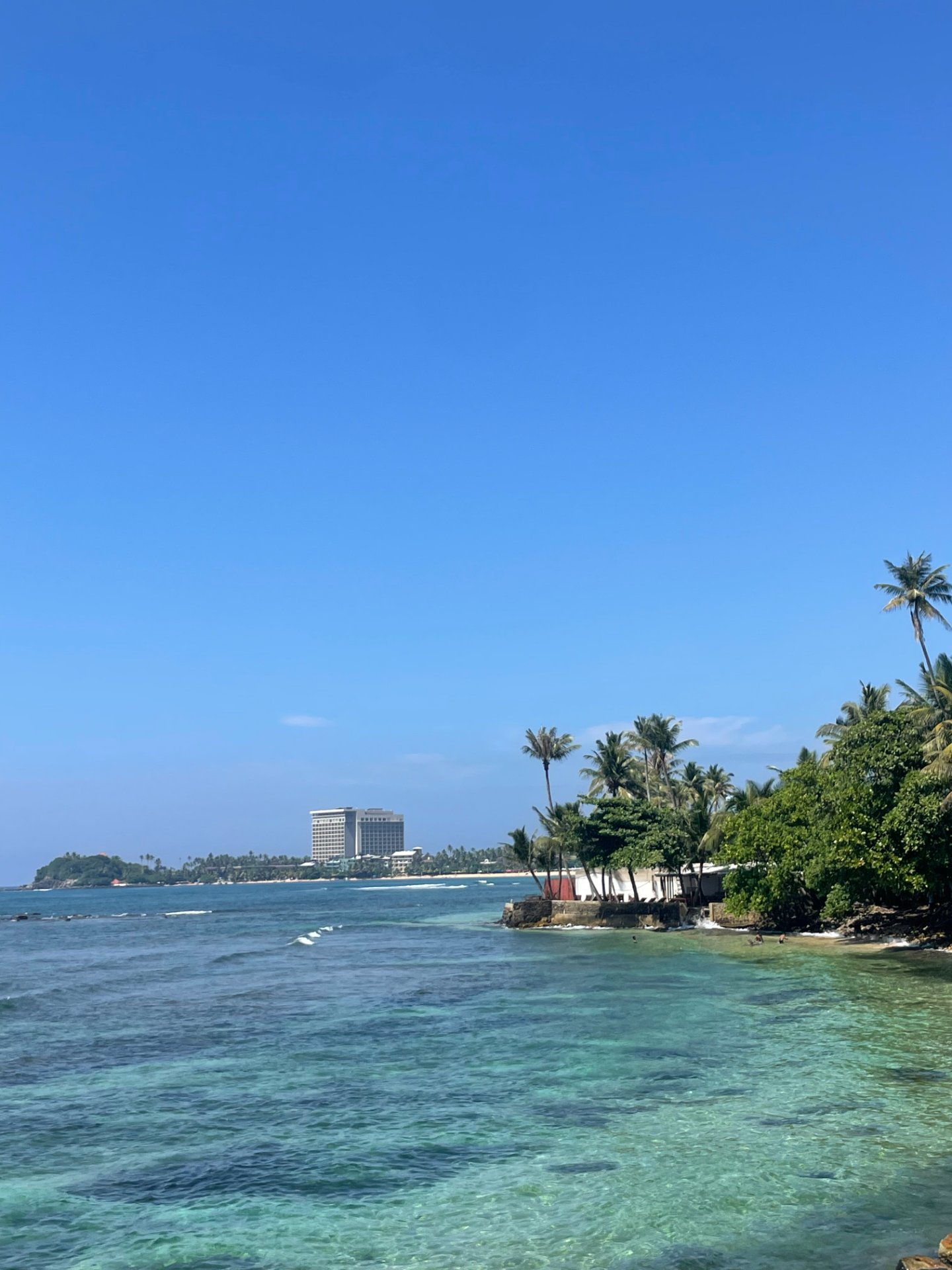 Expansive coastal view of the turquoise Indian Ocean with palm trees and a distant building under a clear blue sky in Sri Lanka.