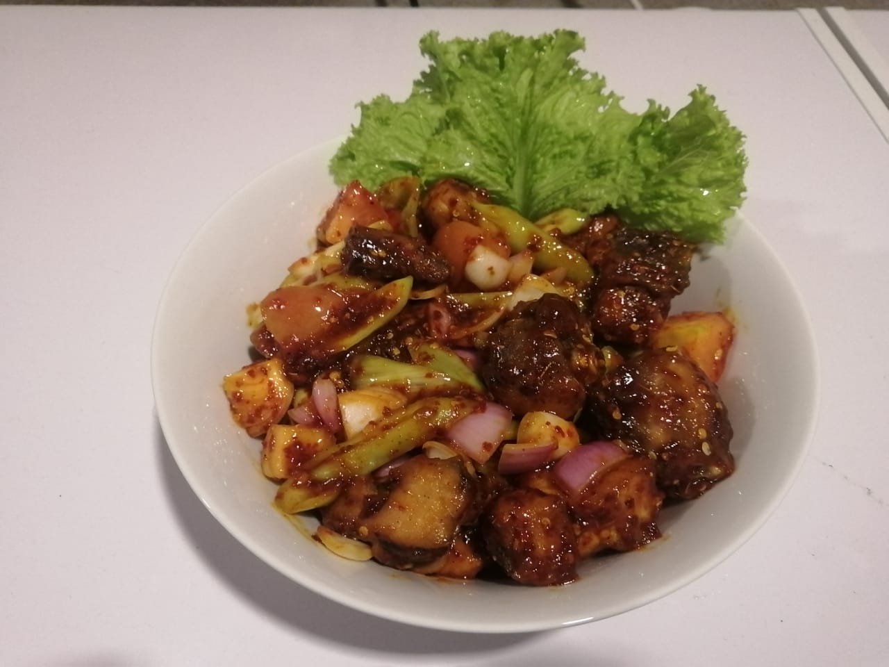 A close-up of a vibrant, saucy Sri Lankan dish with vegetables and meat, garnished with fresh lettuce in a white bowl.