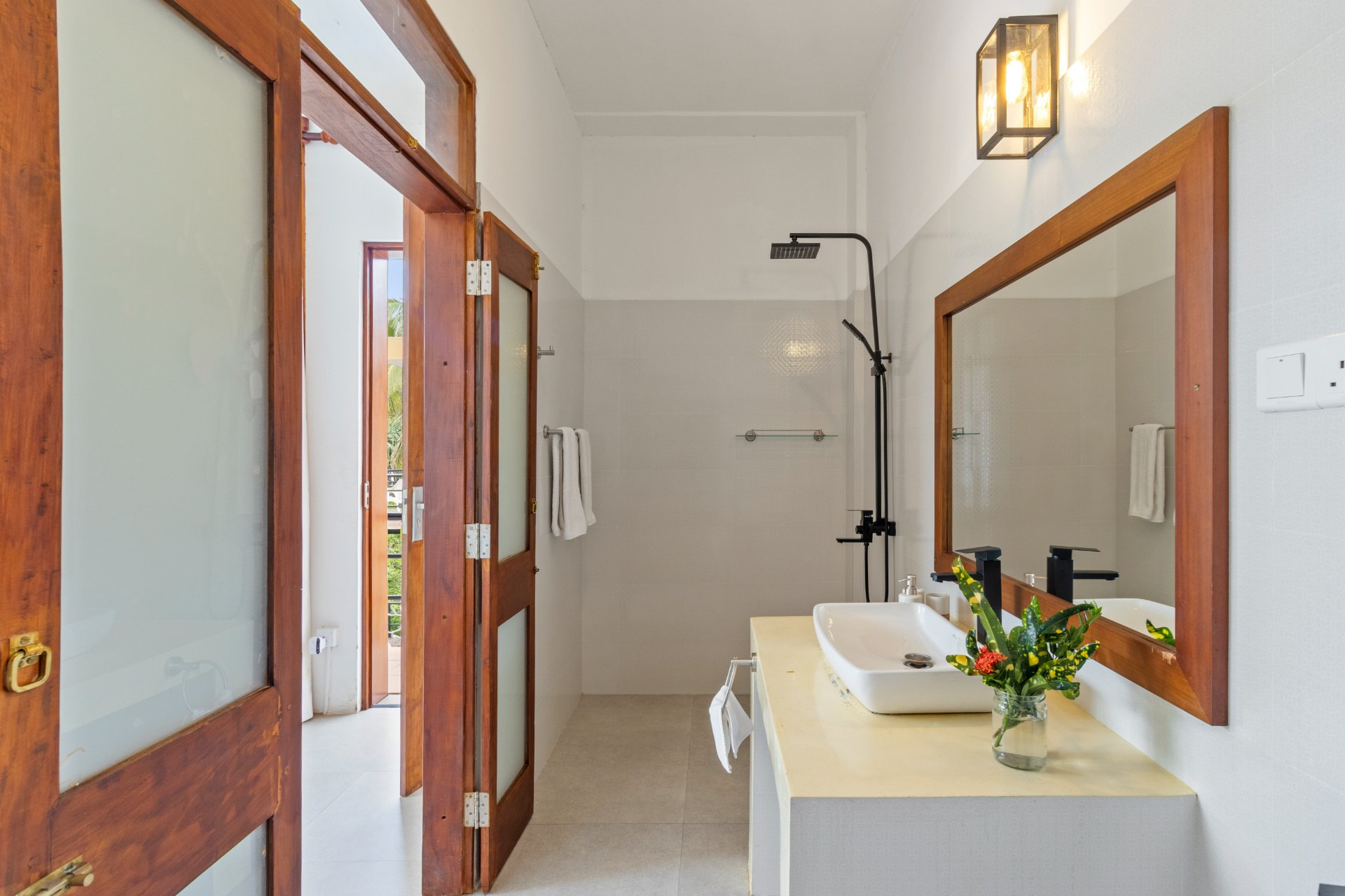 A modern, bright bathroom at Ko Lake Villa with a white basin, wooden mirror, black shower, and open doors revealing green foliage.