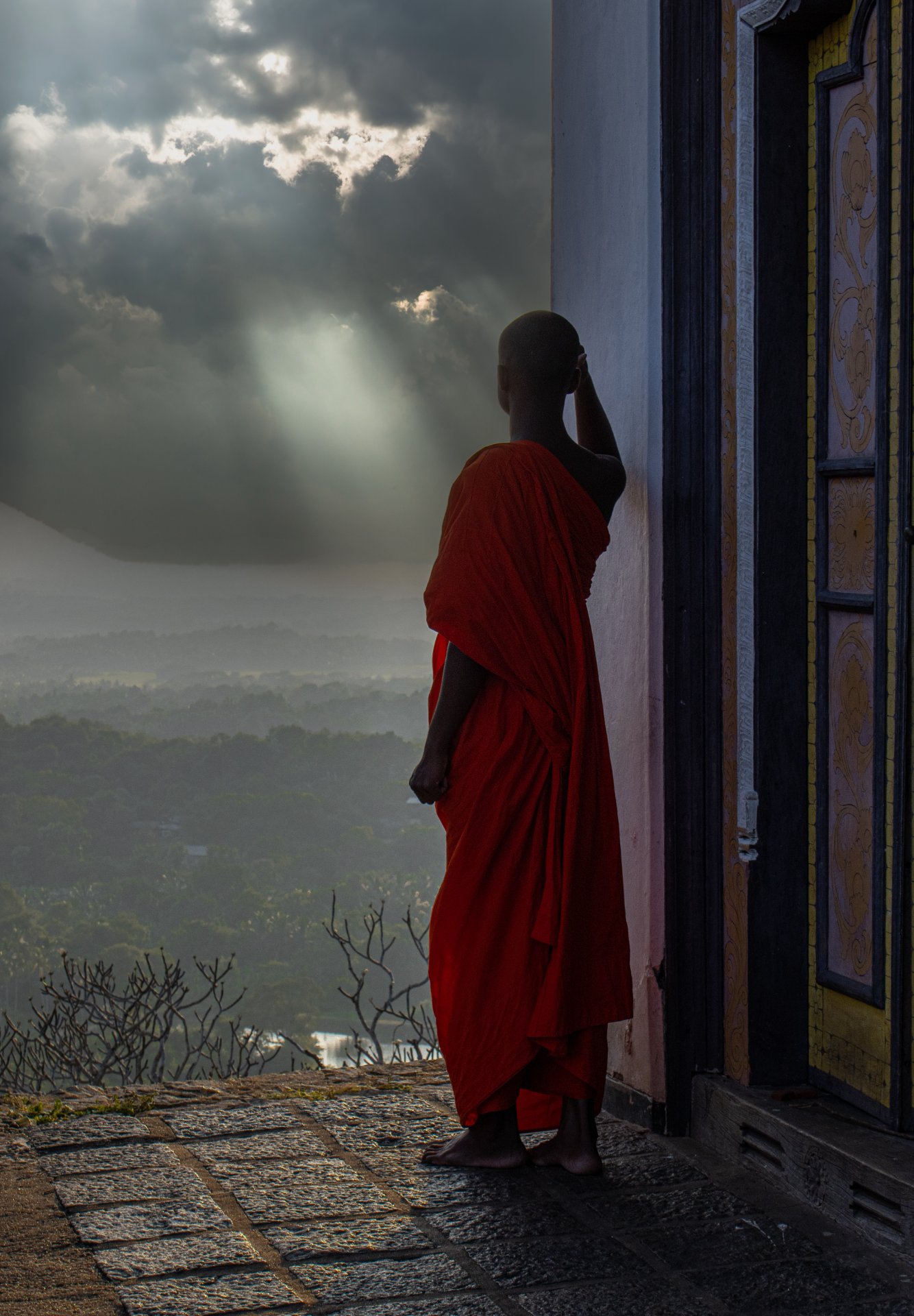 Young Monk Gazing Sri Lanka Landscape - Ko Lake Villa boutique lakefront accommodation on Koggala Lake Sri Lanka: A young Buddhist monk in orange robes looks out over a misty Sri Lankan landscape with rays of light breaking through dramatic clouds. Spiritual journey.