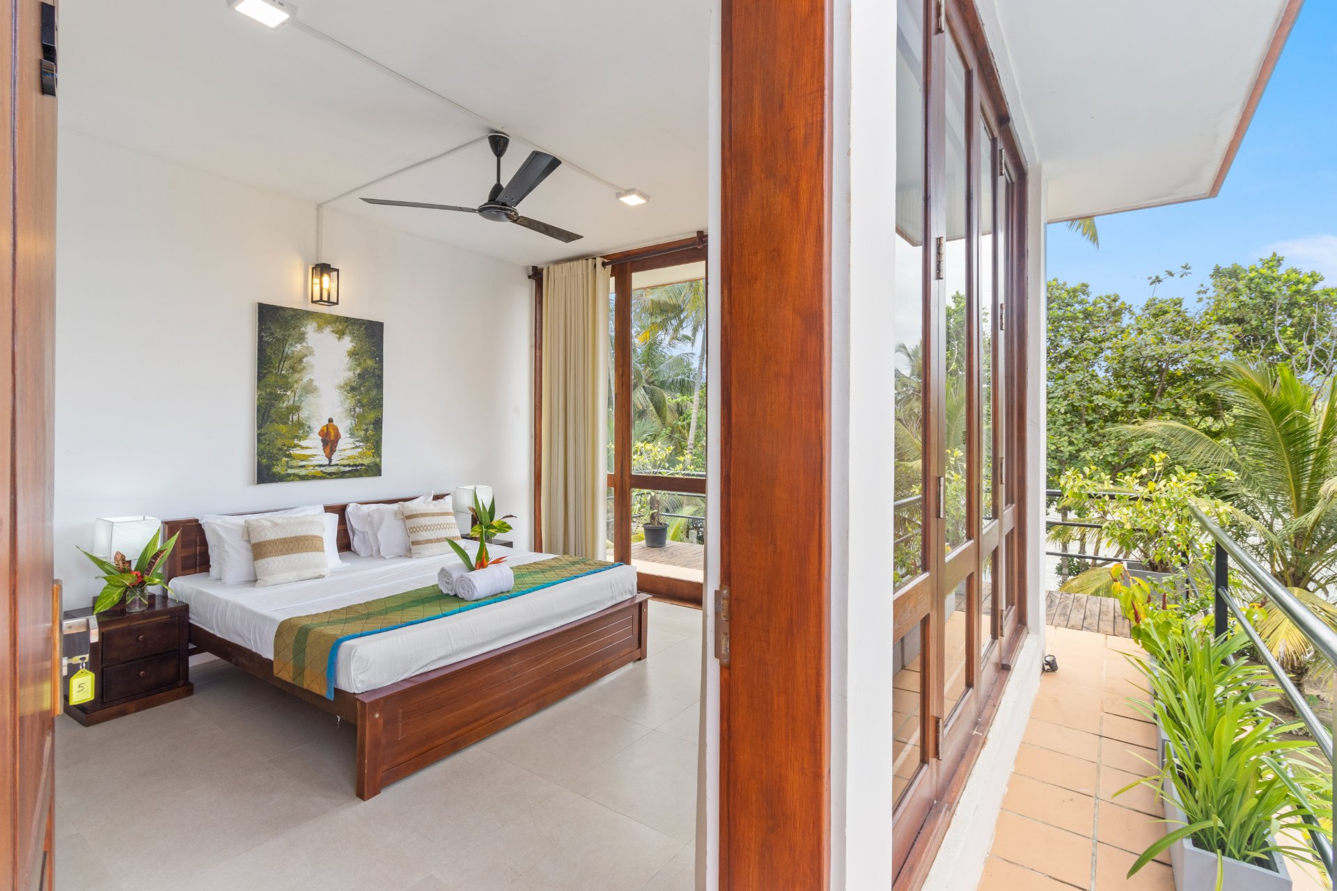 A bright bedroom at Ko Lake Villa with a king-sized bed, white linens, a ceiling fan, and large glass doors opening to a balcony with lush tropical views and a glimpse of the lake.