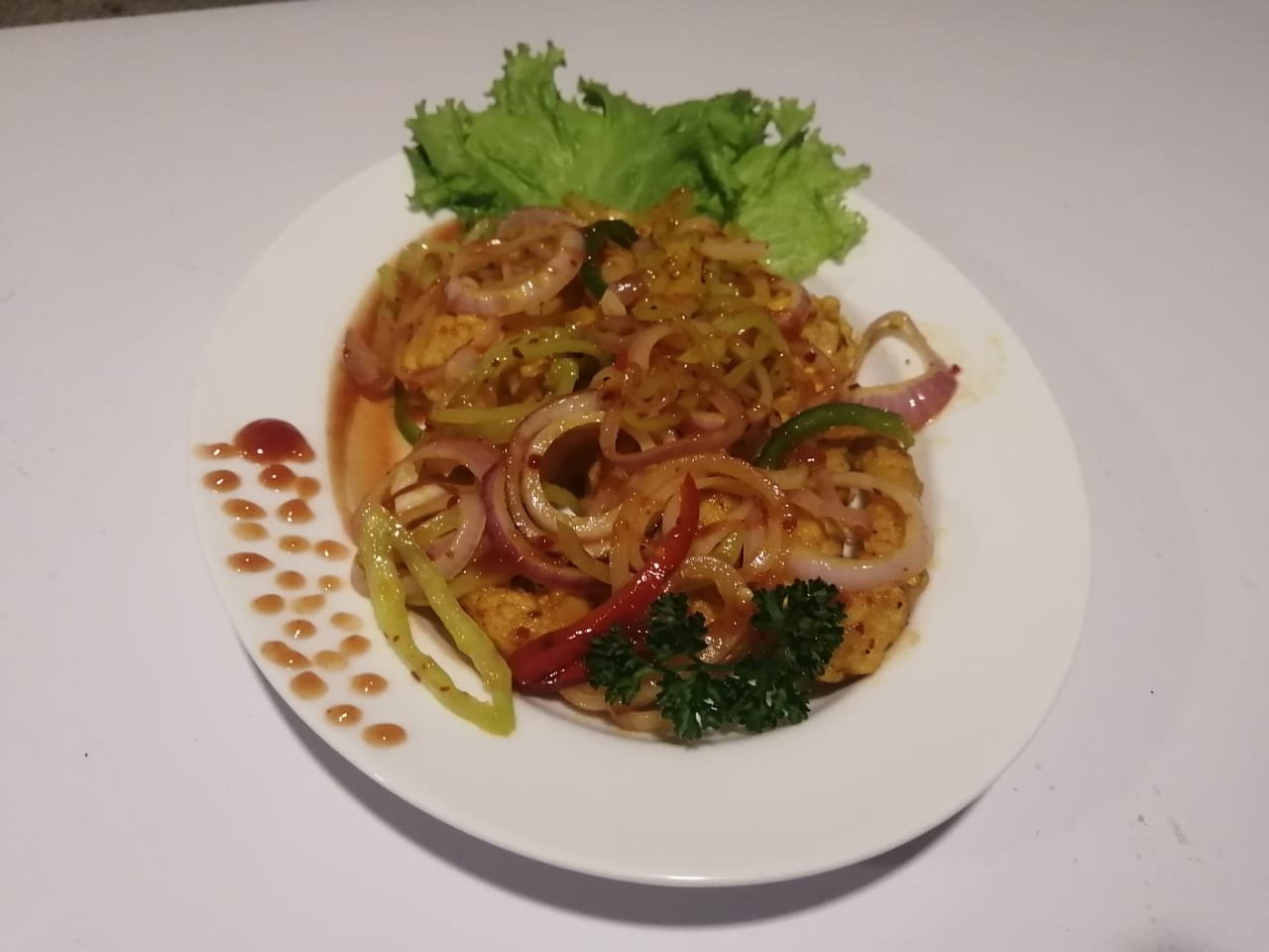 A gourmet dish featuring fried protein, topped with sautéed red onion rings, green bell pepper strips, and red chilies, garnished with lettuce and parsley, on a white plate.