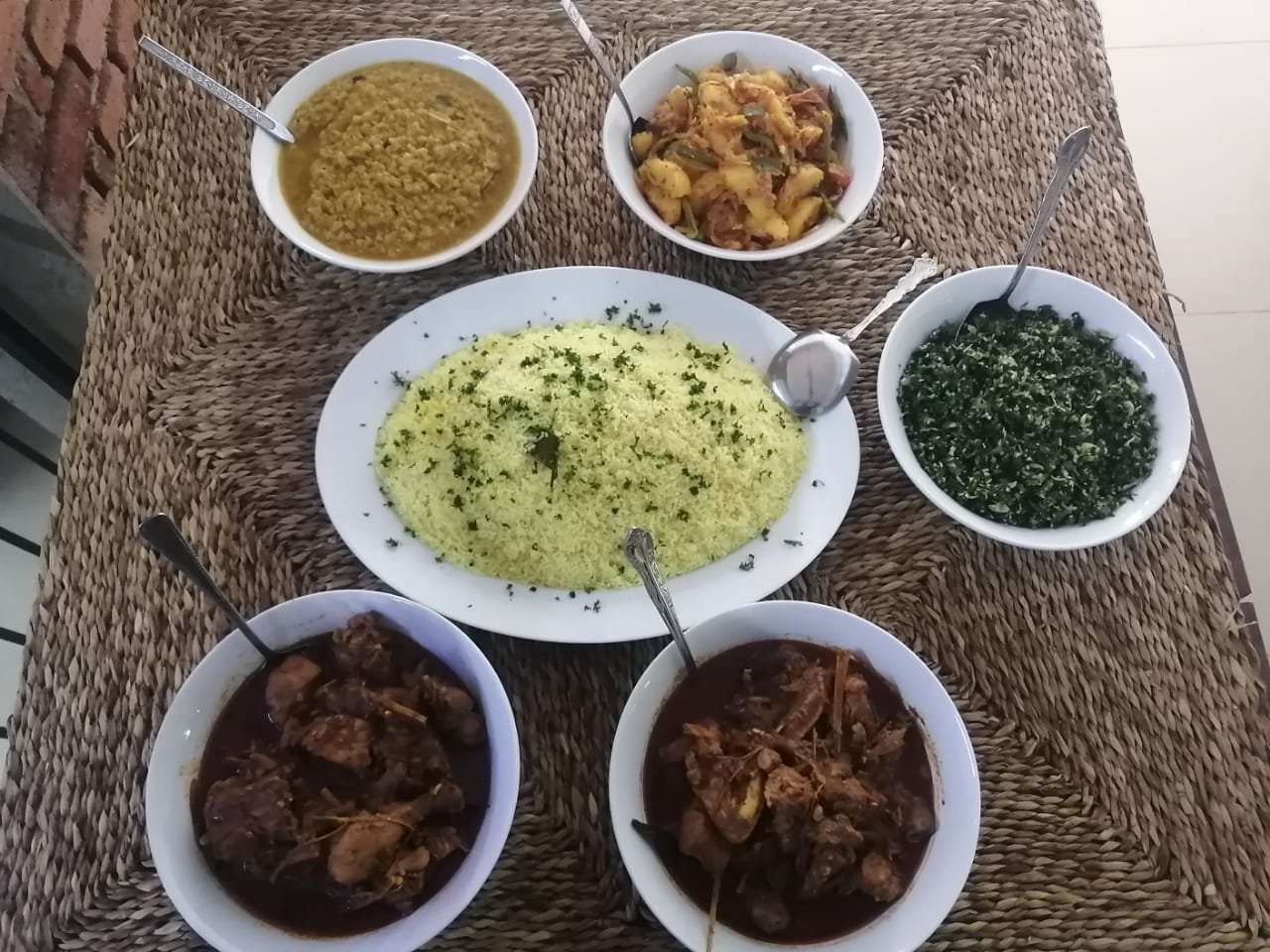 A spread of traditional Sri Lankan dishes including yellow rice, dhal, chicken curries, potatoes, and gotu kola sambol on a woven placemat.
