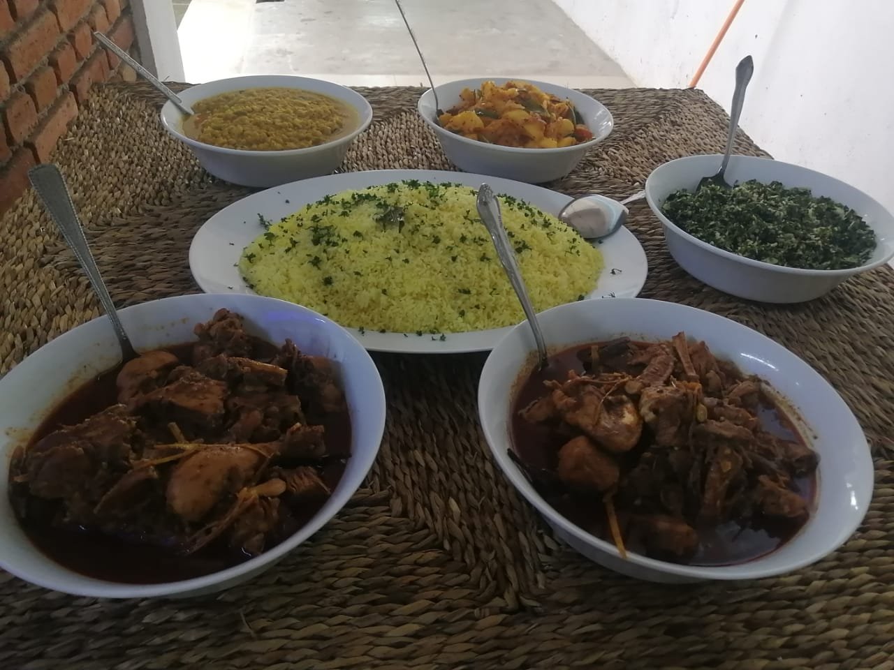 A spread of traditional Sri Lankan dishes, including two bowls of chicken curry, yellow rice, dhal, and vegetable sides, on a woven mat.