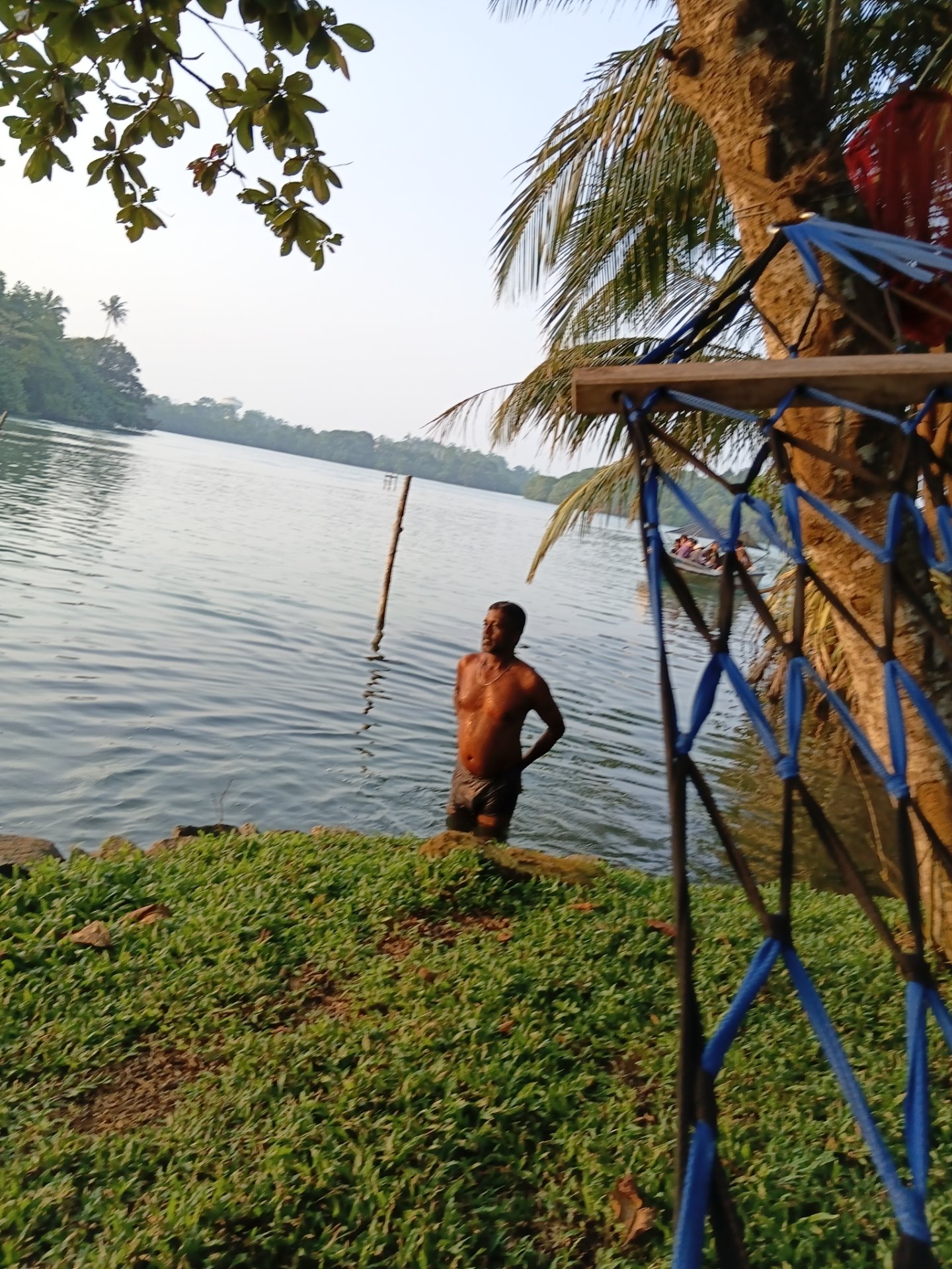 Man wading in clear Koggala Lake, Sri Lanka, with lush green shore, palm trees, and distant boat. A vibrant blue and black hammock hangs nearby.