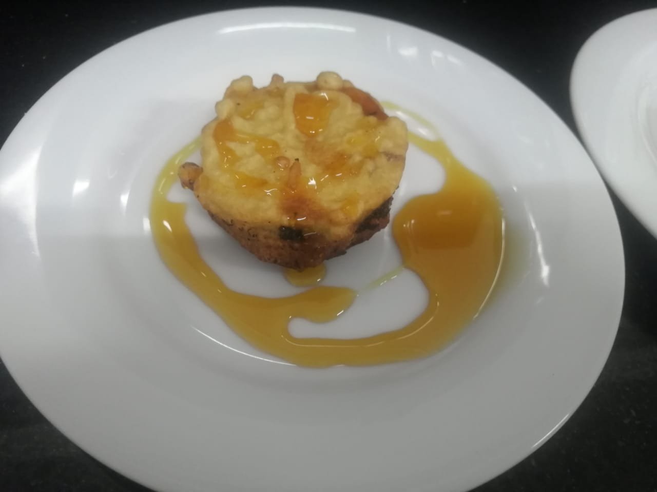 Close-up of a golden-brown dessert, possibly a waffle or fritter, drizzled with glossy amber syrup on a white plate.
