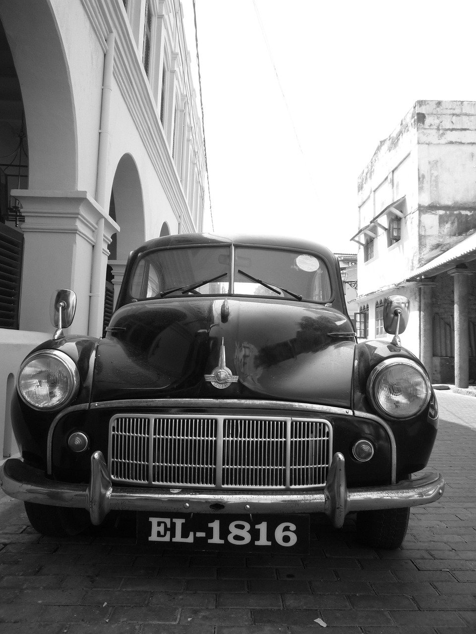 A classic black Morris Minor car, EL-1816, in black and white, parked on a cobblestone street in front of colonial-style buildings in Galle.