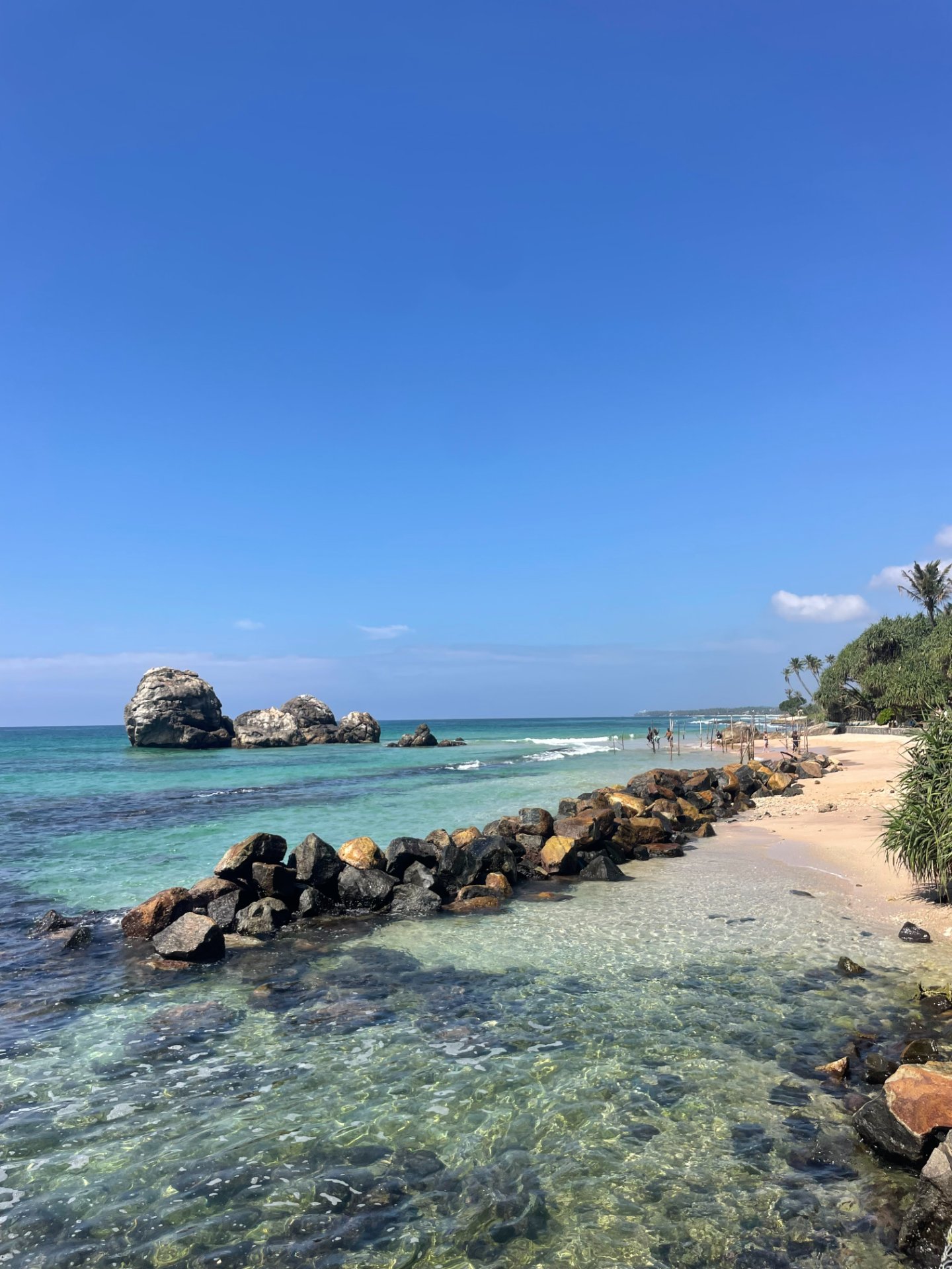 Sunny Koggala Beach in Sri Lanka with clear turquoise water, sandy shore, offshore rocks, and a rocky breakwater.
